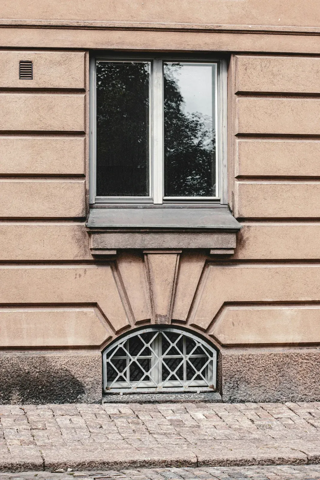 A rectangular window sits above a smaller, arched, grated cellar window on a tan building facade with horizontal lines.