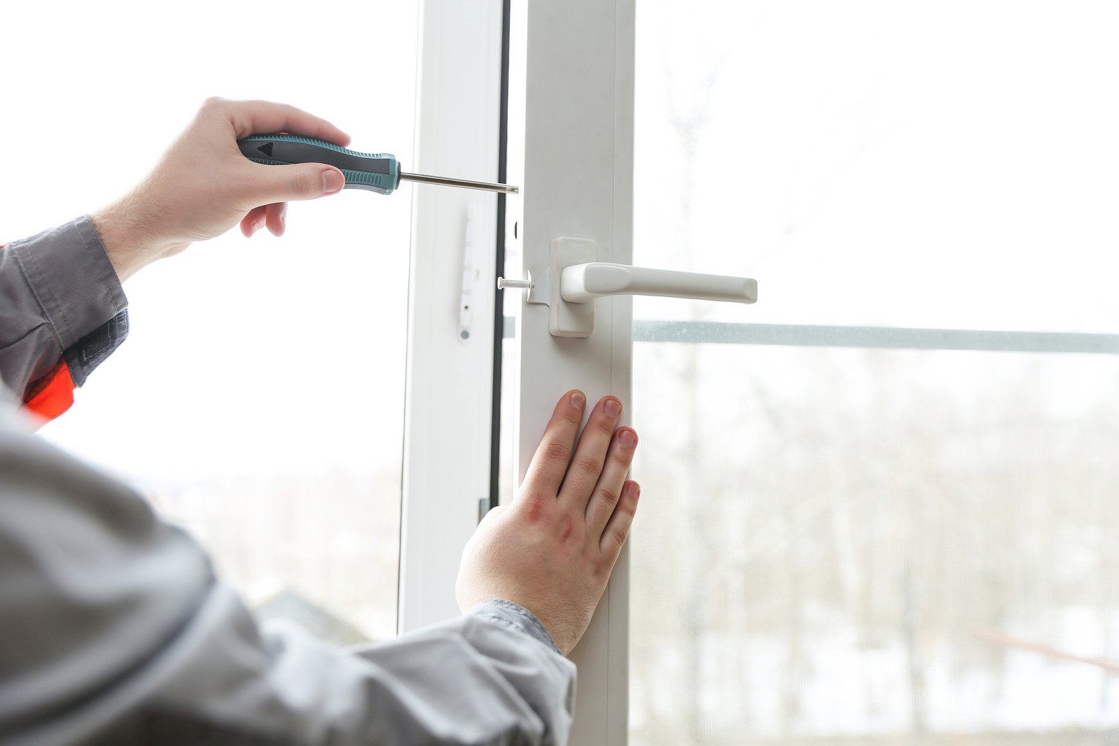 A person in a gray uniform uses a screwdriver to repair a white window frame near a handle.