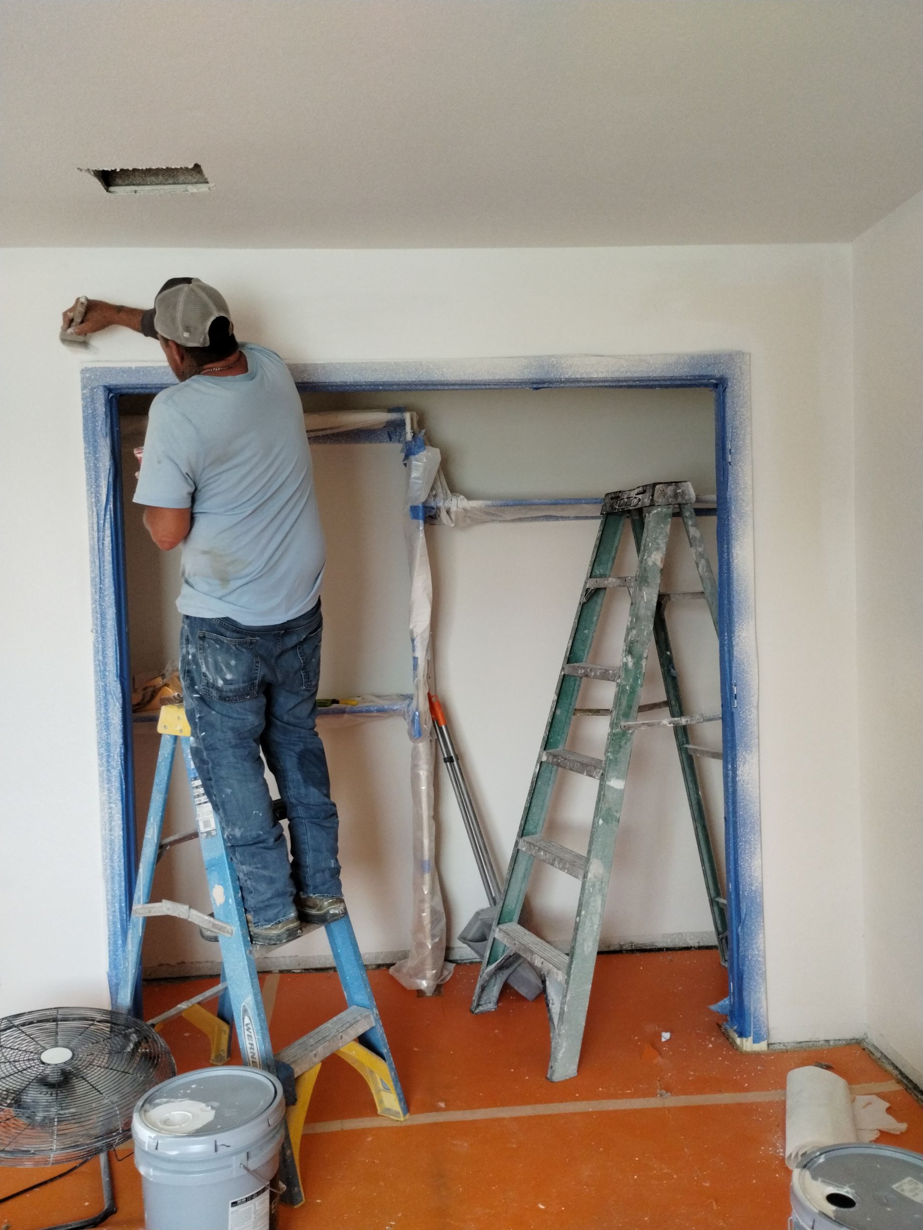 Person painting trim around a doorway while standing on a ladder. Blue painter's tape, paint cans, and another ladder are also visible.