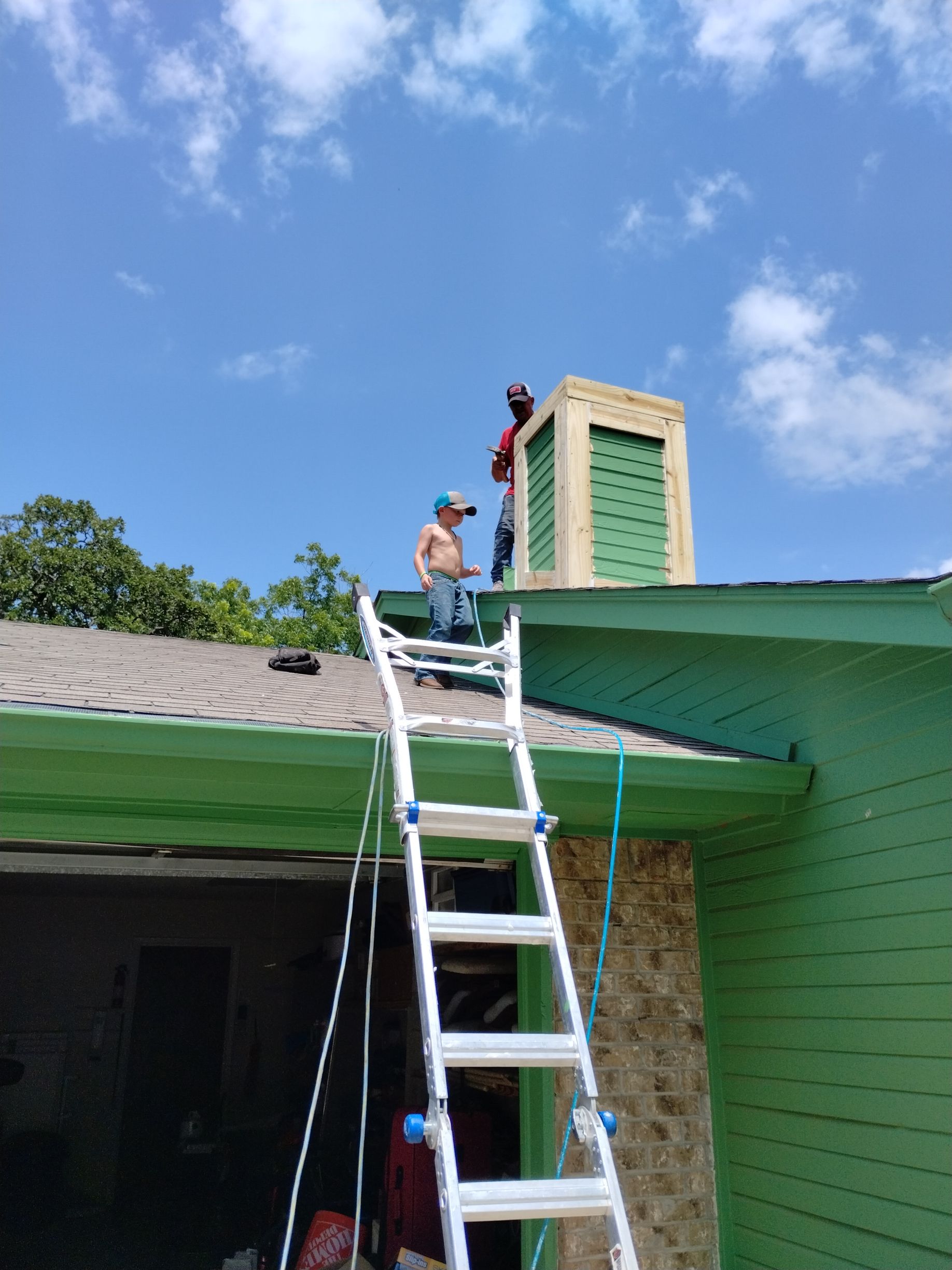 Two men on a roof, one on a ladder, working on a green chimney under a blue sky.