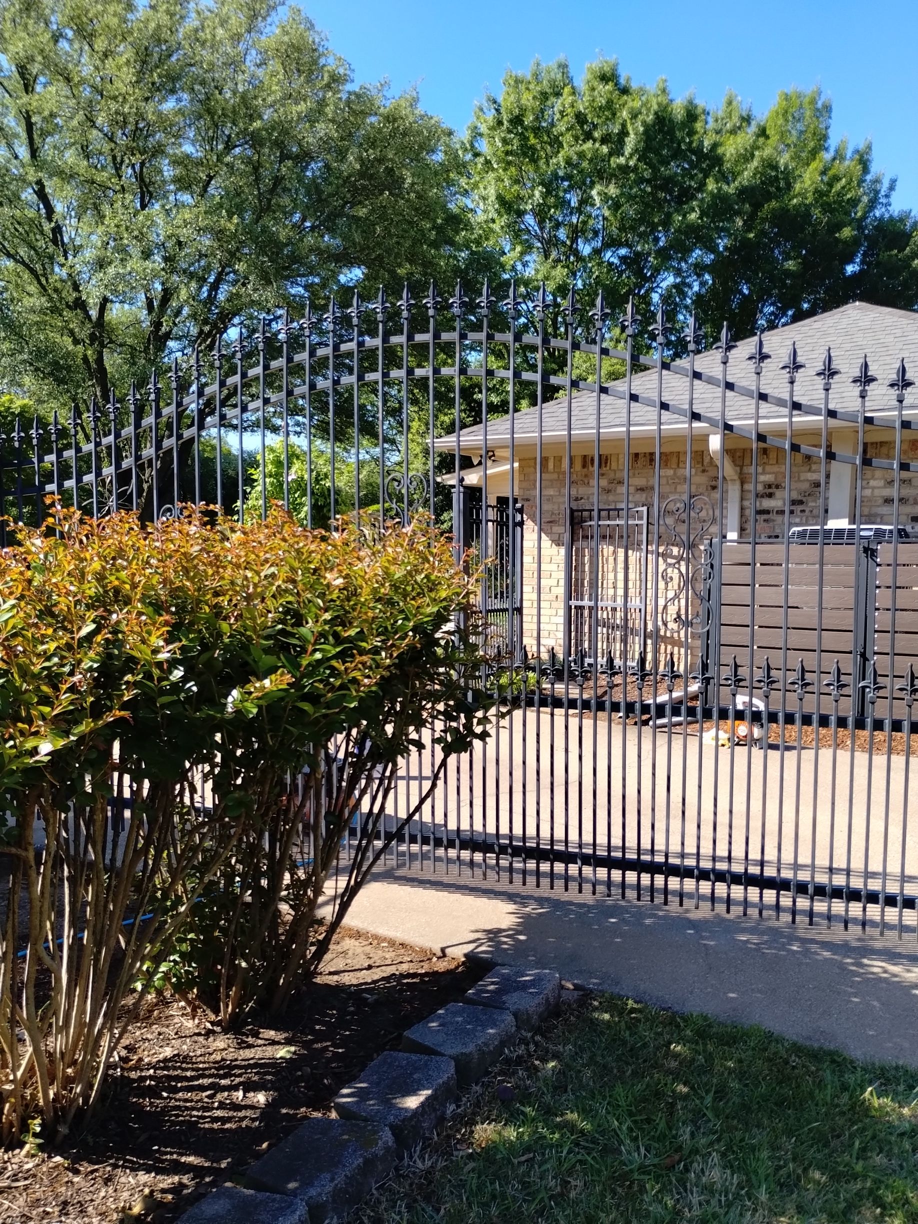 Black iron fence in front of a beige brick house, shrubbery in foreground, blue sky.