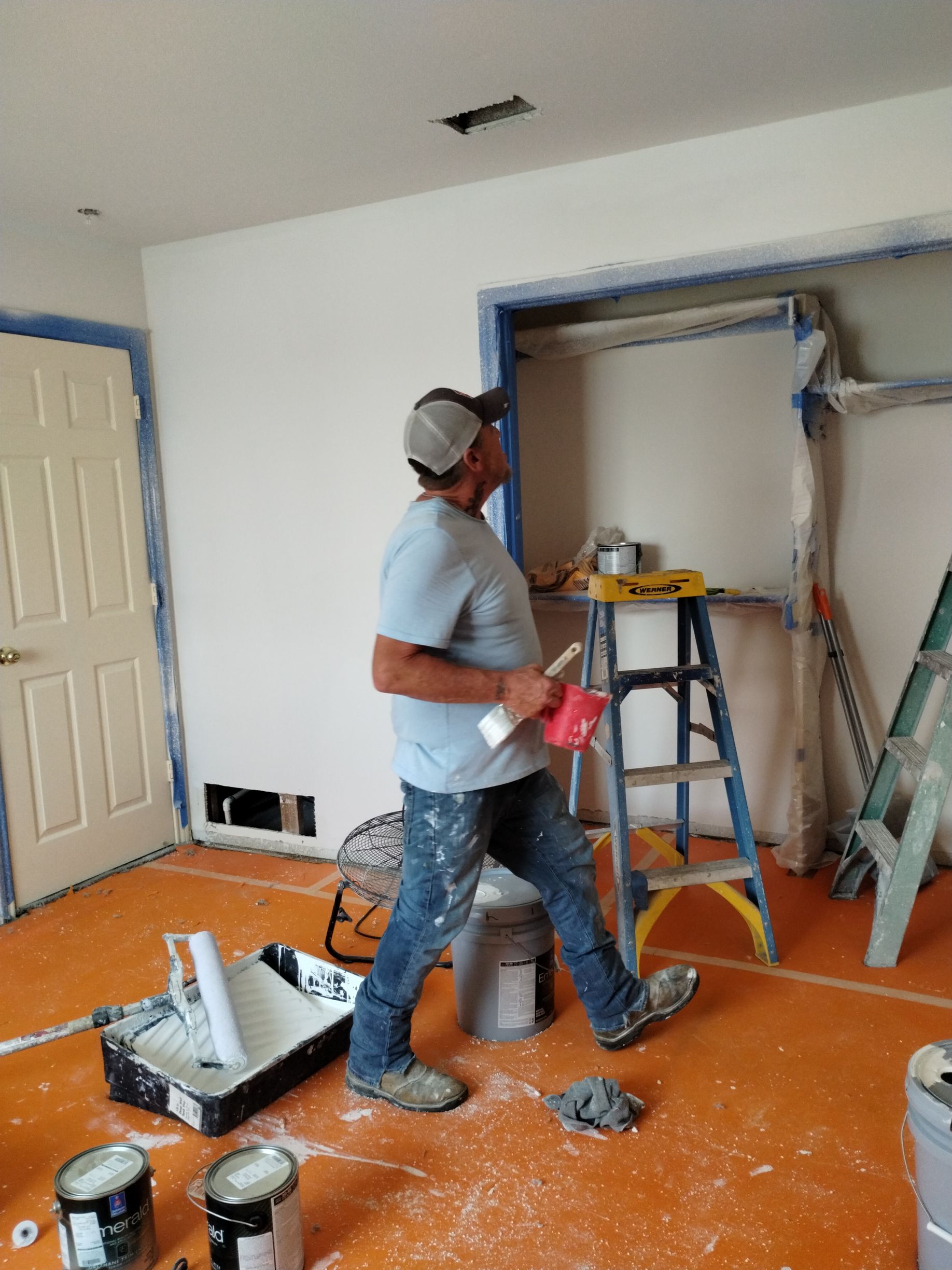 Man in paint-splattered jeans and shirt surveys freshly painted room. Masked trim, orange flooring.
