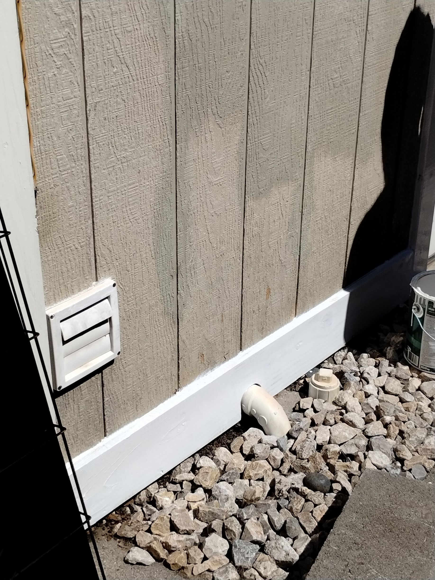 A light brown building with a white dryer vent and drain pipe near a stone ground.