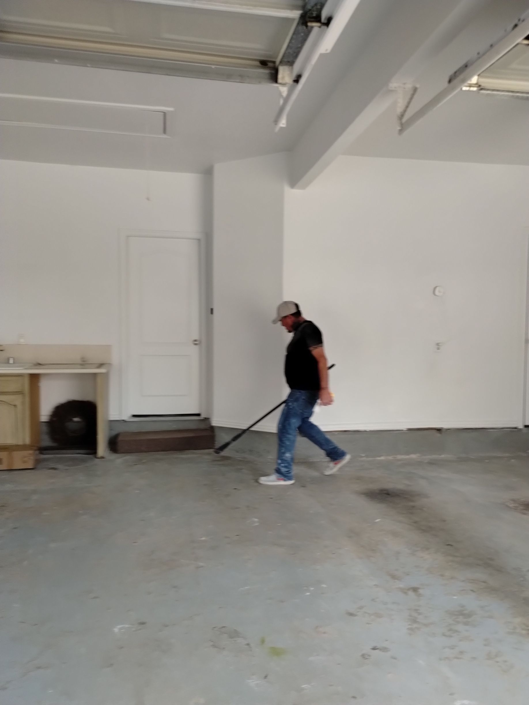Man raking a dirty garage floor. He is wearing a cap, black shirt, and jeans. The walls are white.