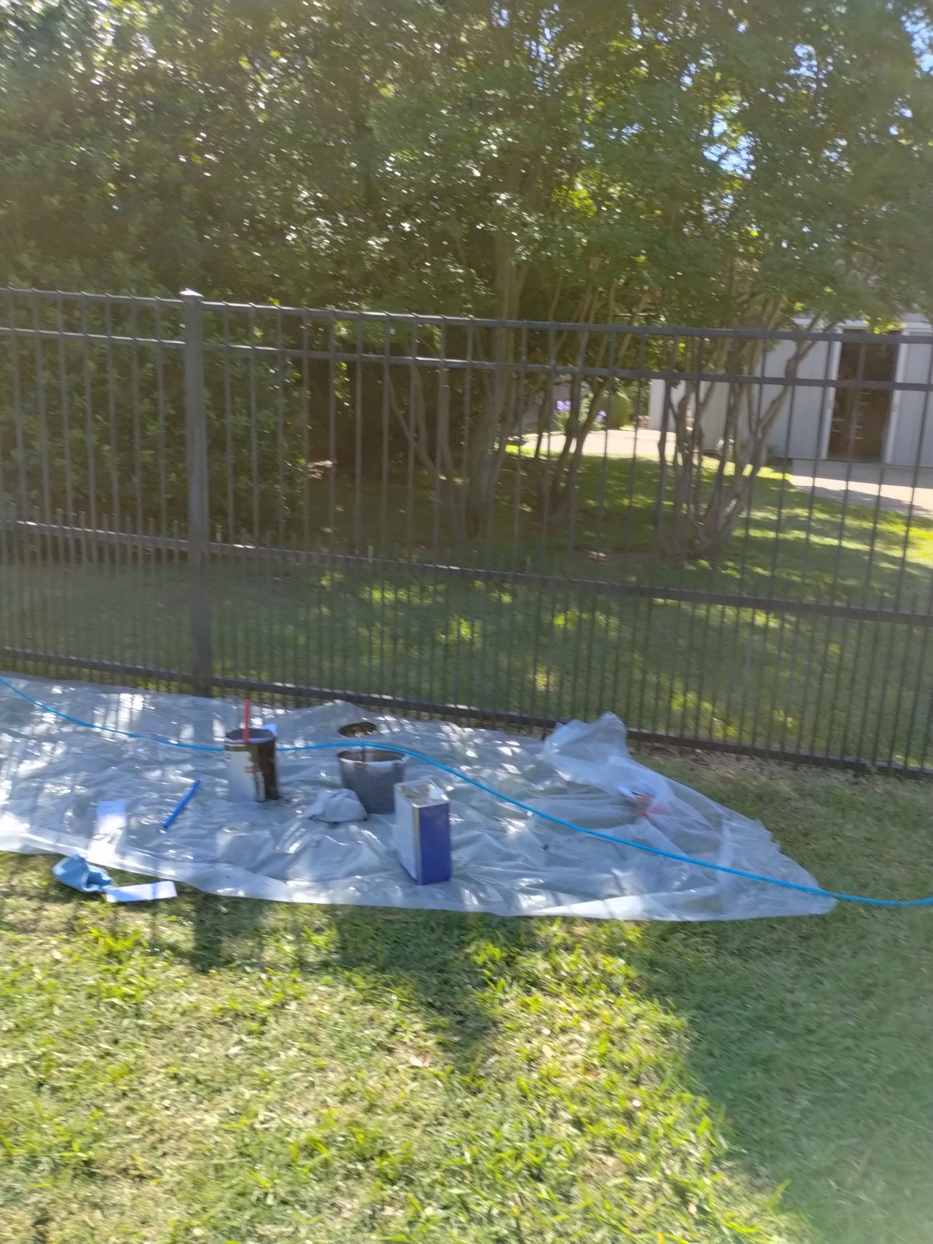 A tarp with painting supplies on grass in front of a black fence and trees.