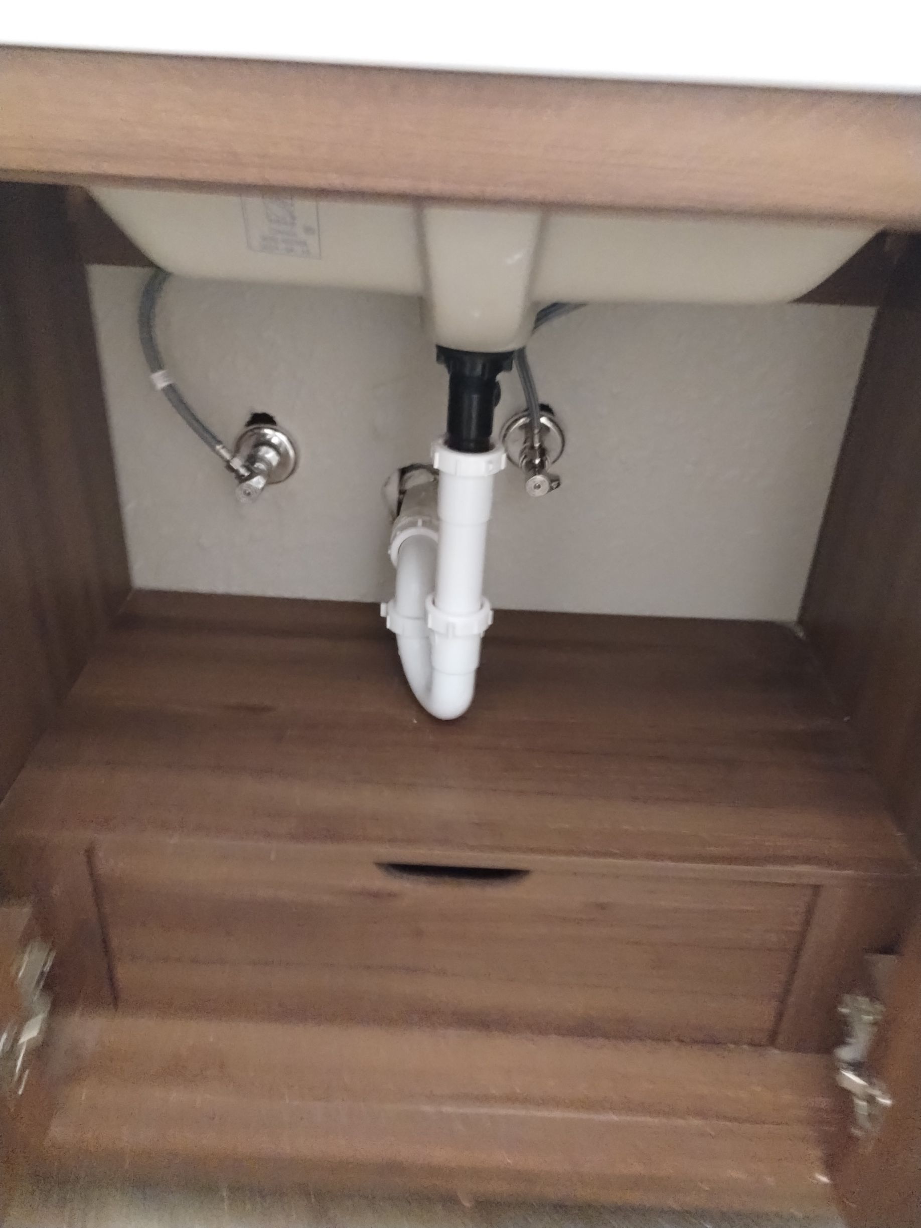 Under-sink cabinet with plumbing. Beige sink, white drain pipe, and metal water lines against a brown cabinet.