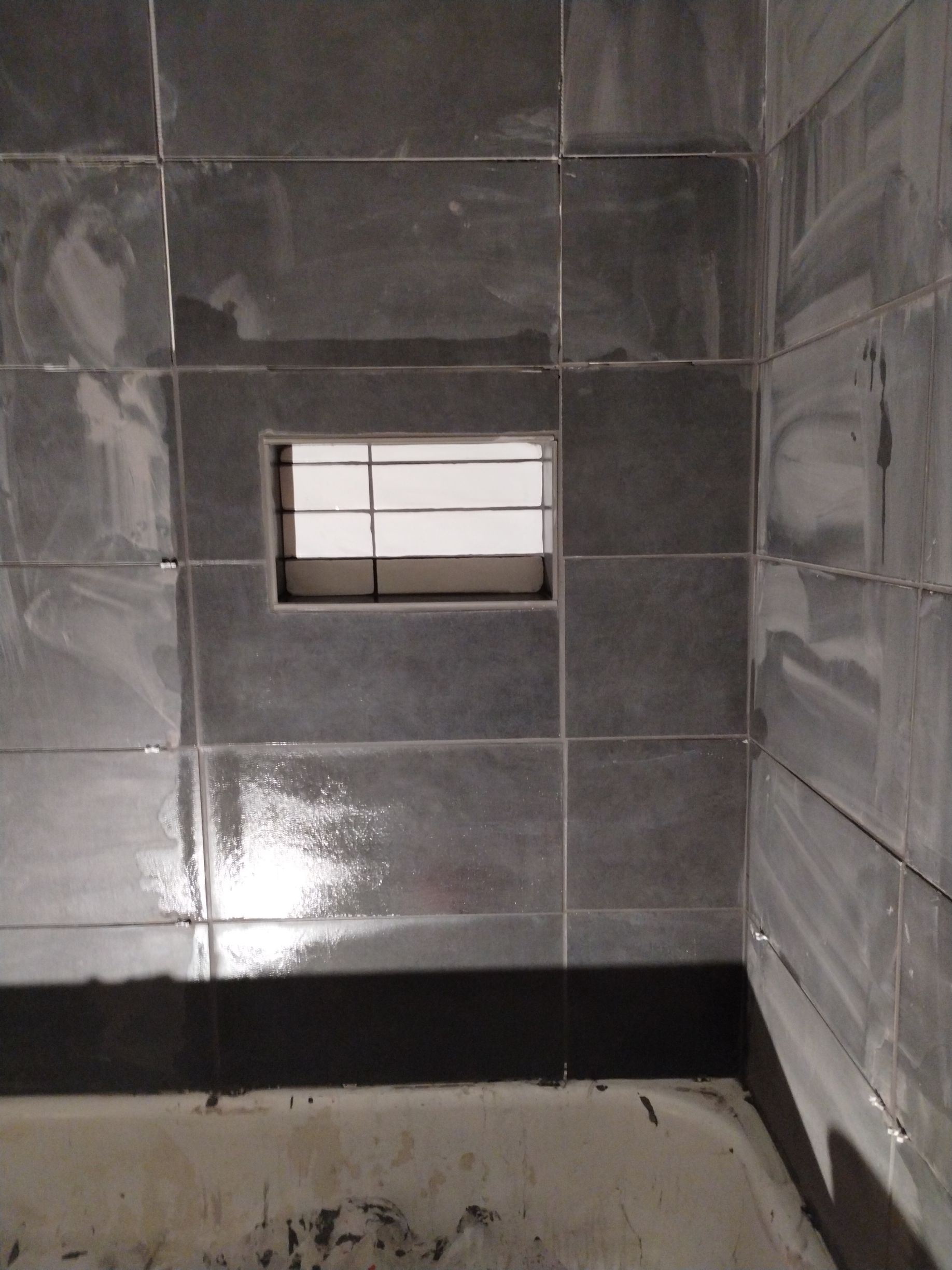 Gray tiled shower with a small window, and a dark baseboard.
