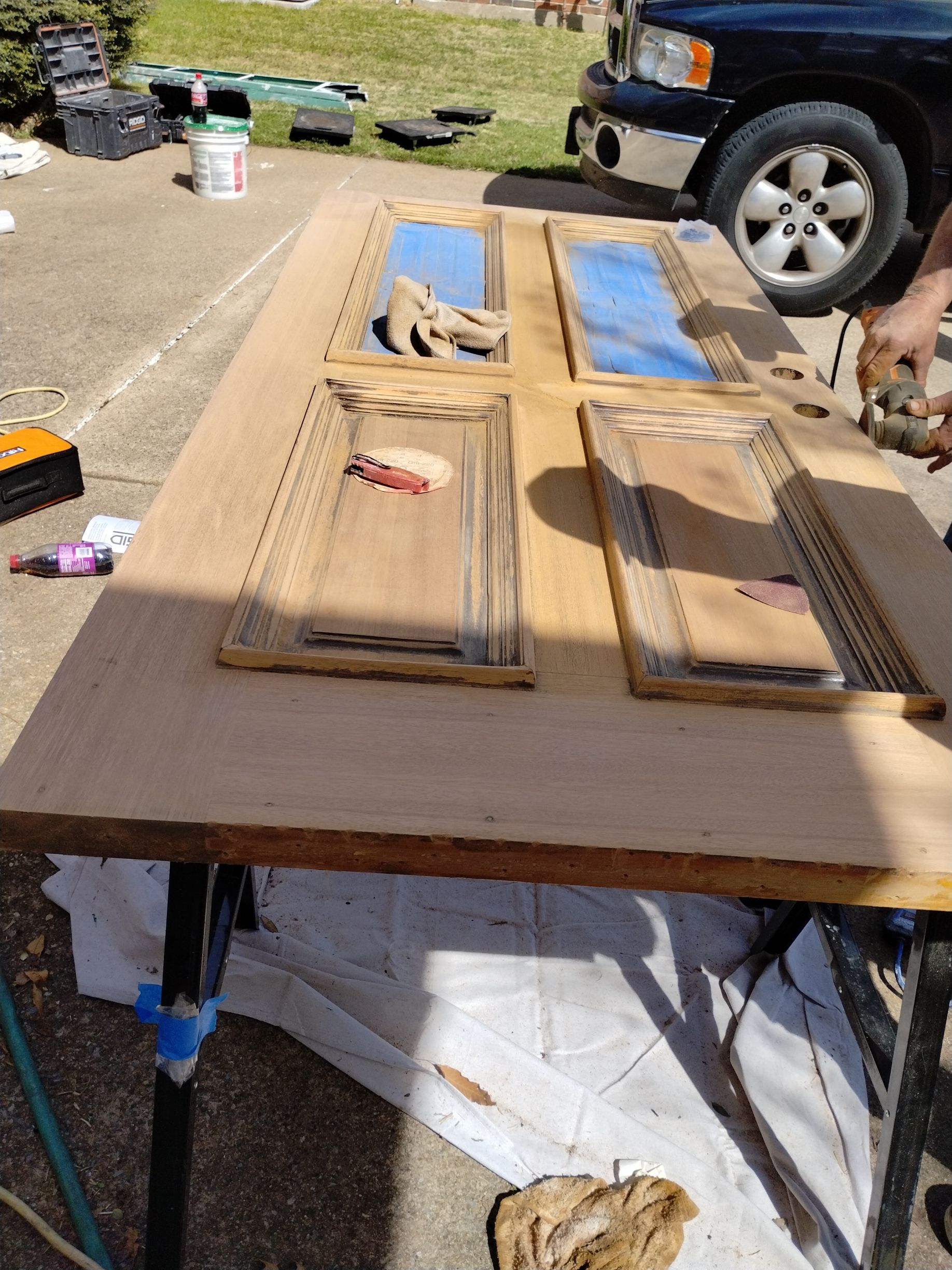 A wooden door laid on a table is being sanded outside. Blue tape protects glass panels.