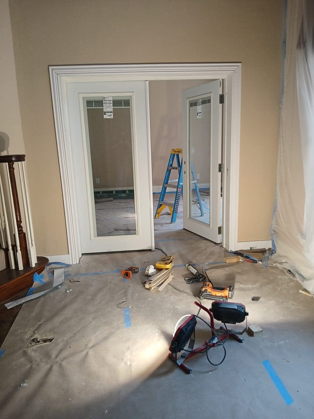 Interior room under renovation with glass doors, tools on the floor, and a ladder in the background.