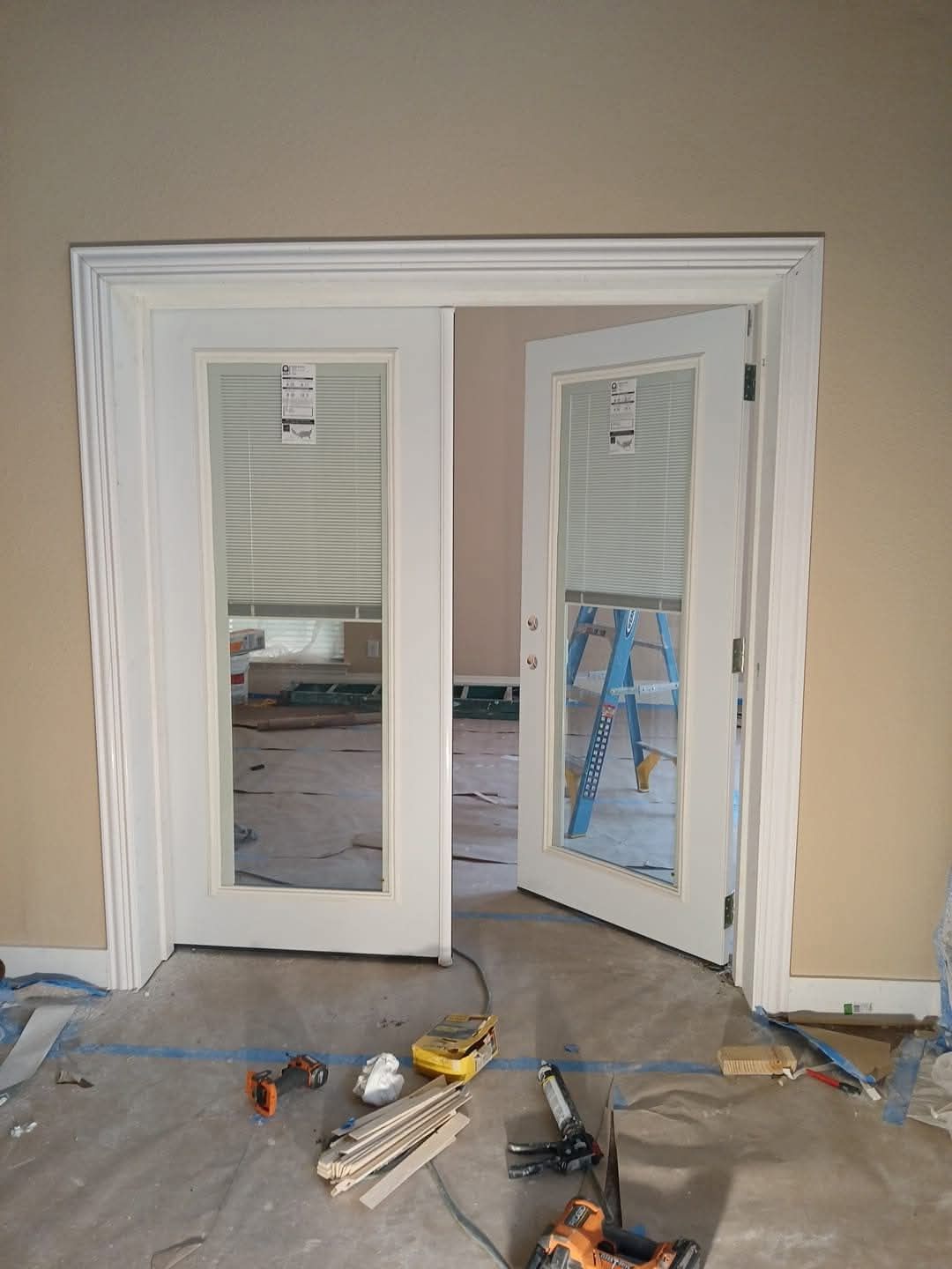 White French doors, one open, with white trim, set in a tan-walled room under construction, tools on floor.