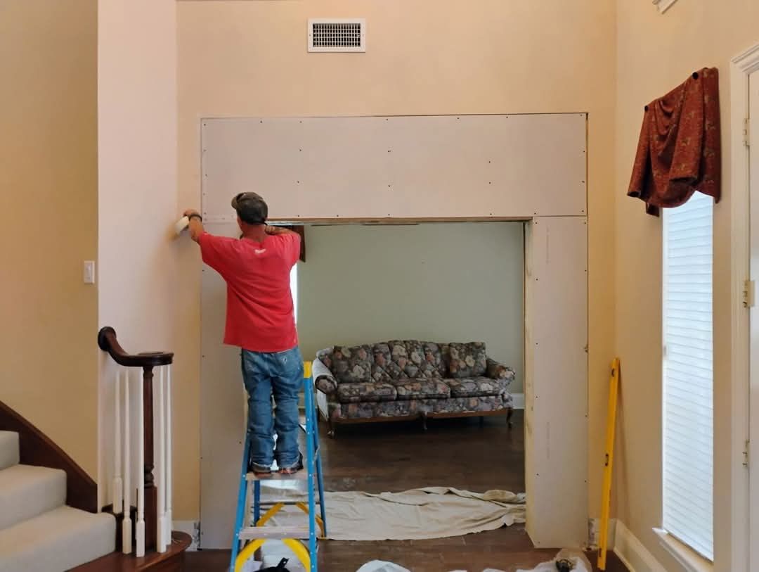 Man on ladder installing drywall in doorway. Beige walls, green room visible, sofa, red shirt, blue jeans.