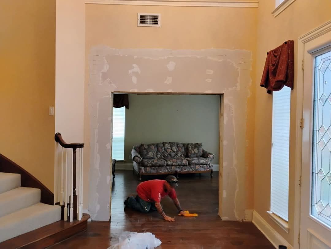 Person cleaning a doorway, home interior, light walls, stairs on the left, sofa visible through doorway.