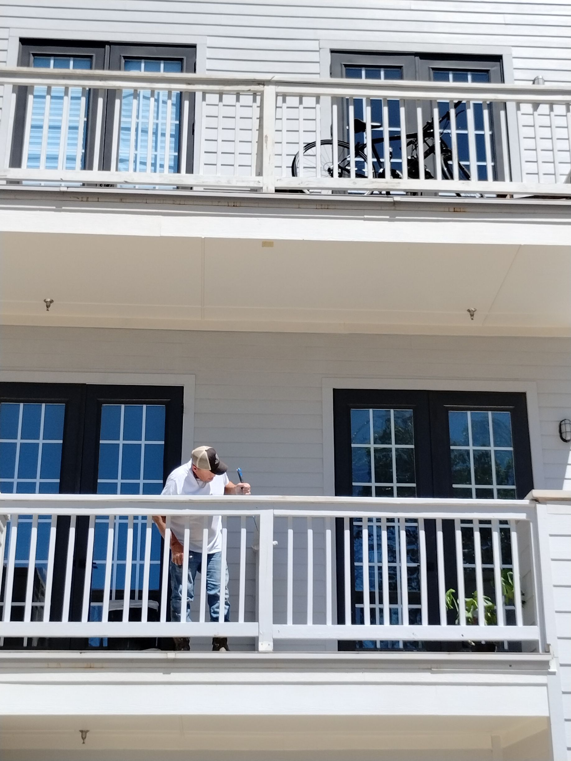 Man on white balcony looking down. White building, blue windows, black doors.