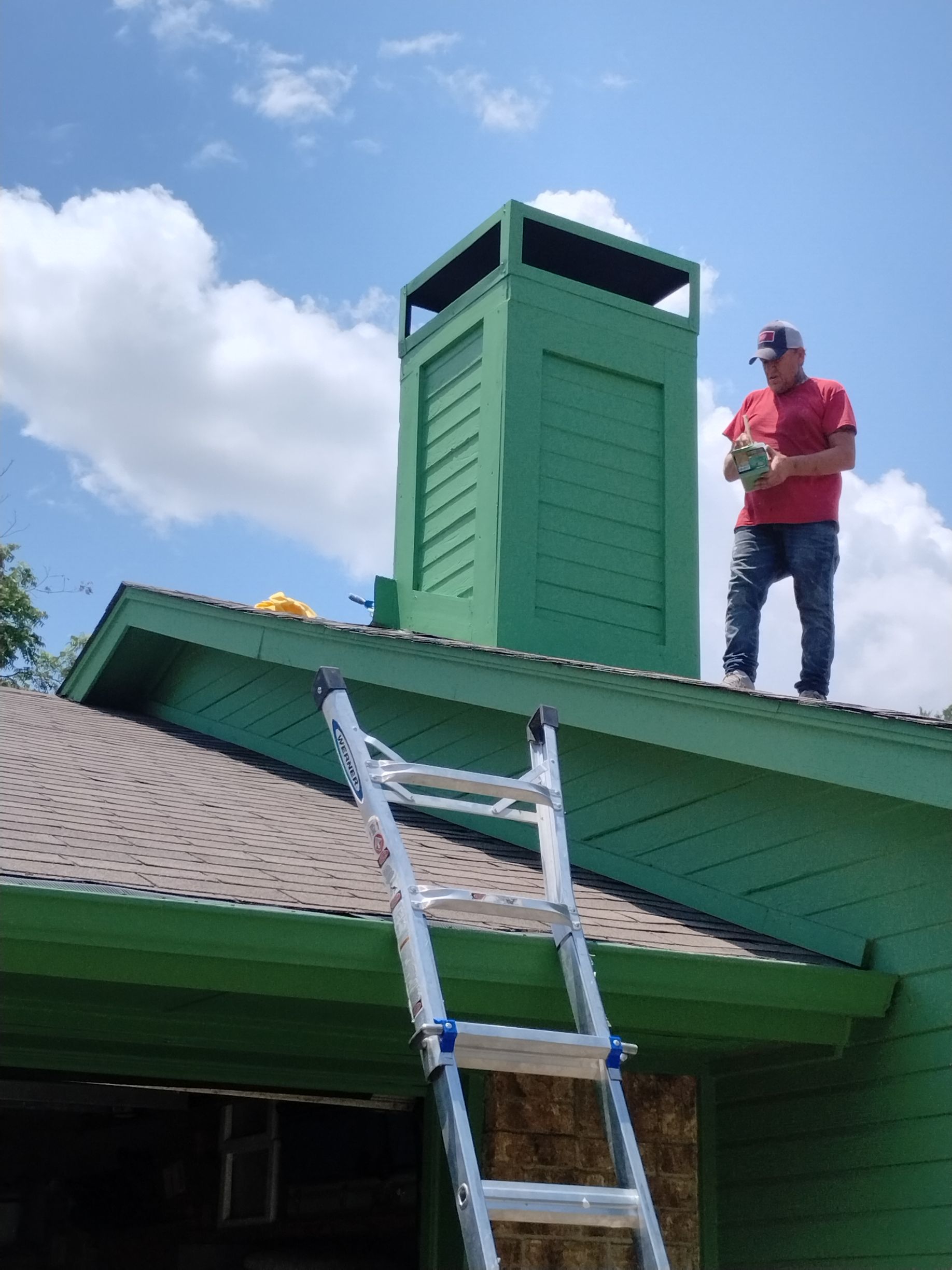 Man on roof near green chimney, holding object. Ladder leads to roof. Sunny, blue sky.