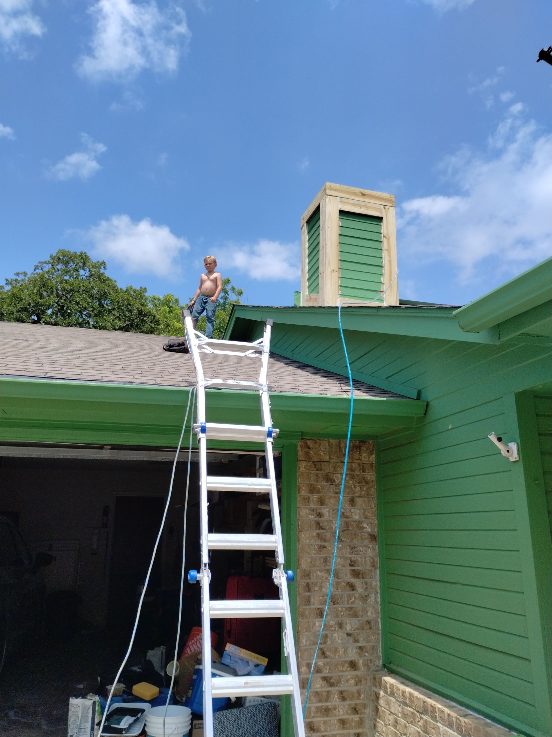 Person on a ladder working on a green roof near a chimney, under a blue sky.