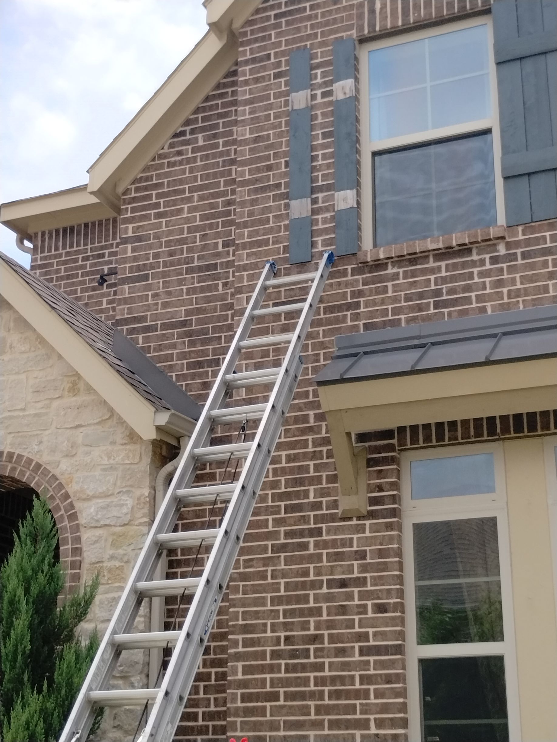Ladder leaning against a brick house, reaching for dark shutters.