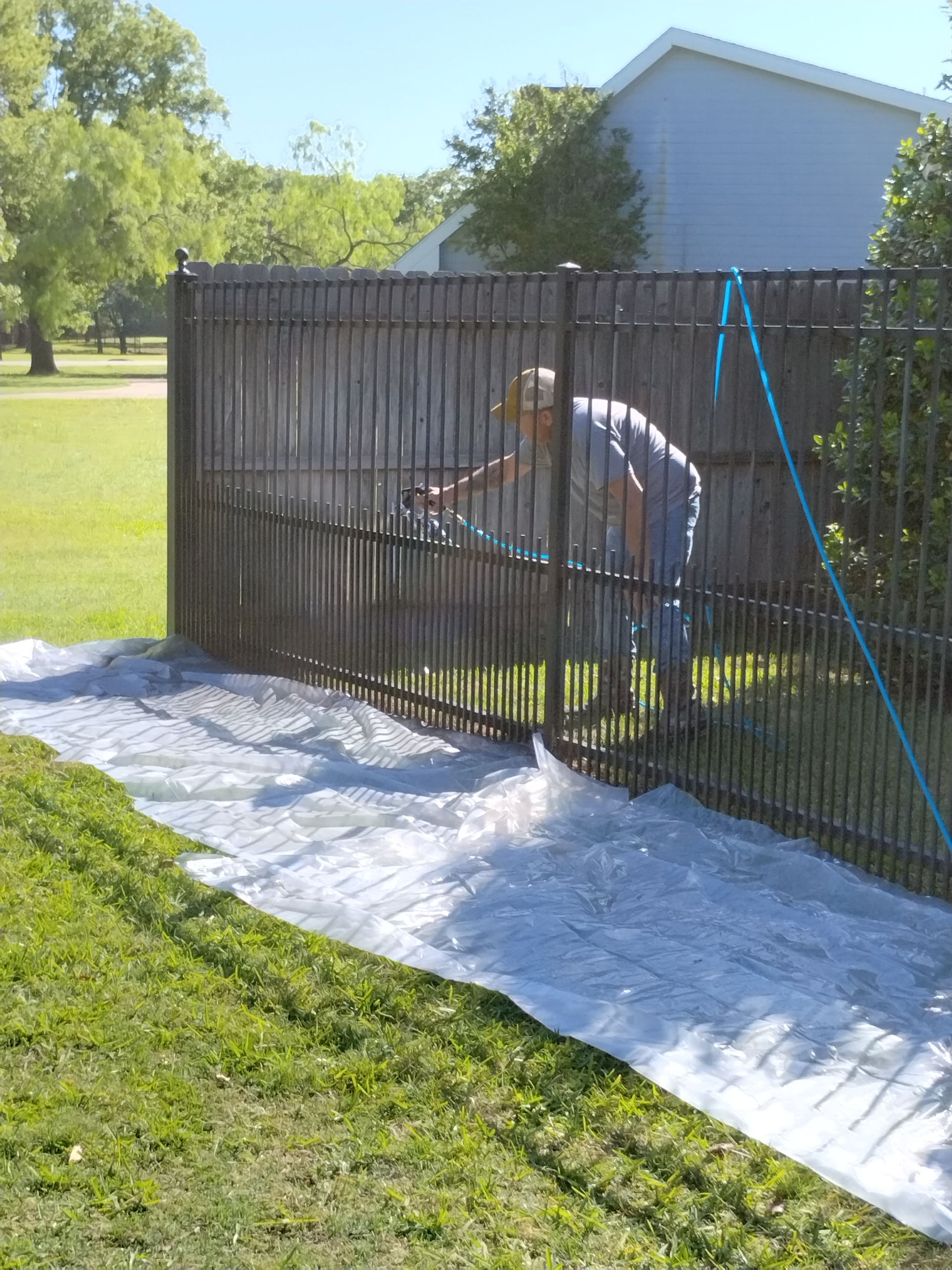 Man spray painting a black fence in a yard, with plastic covering the grass.