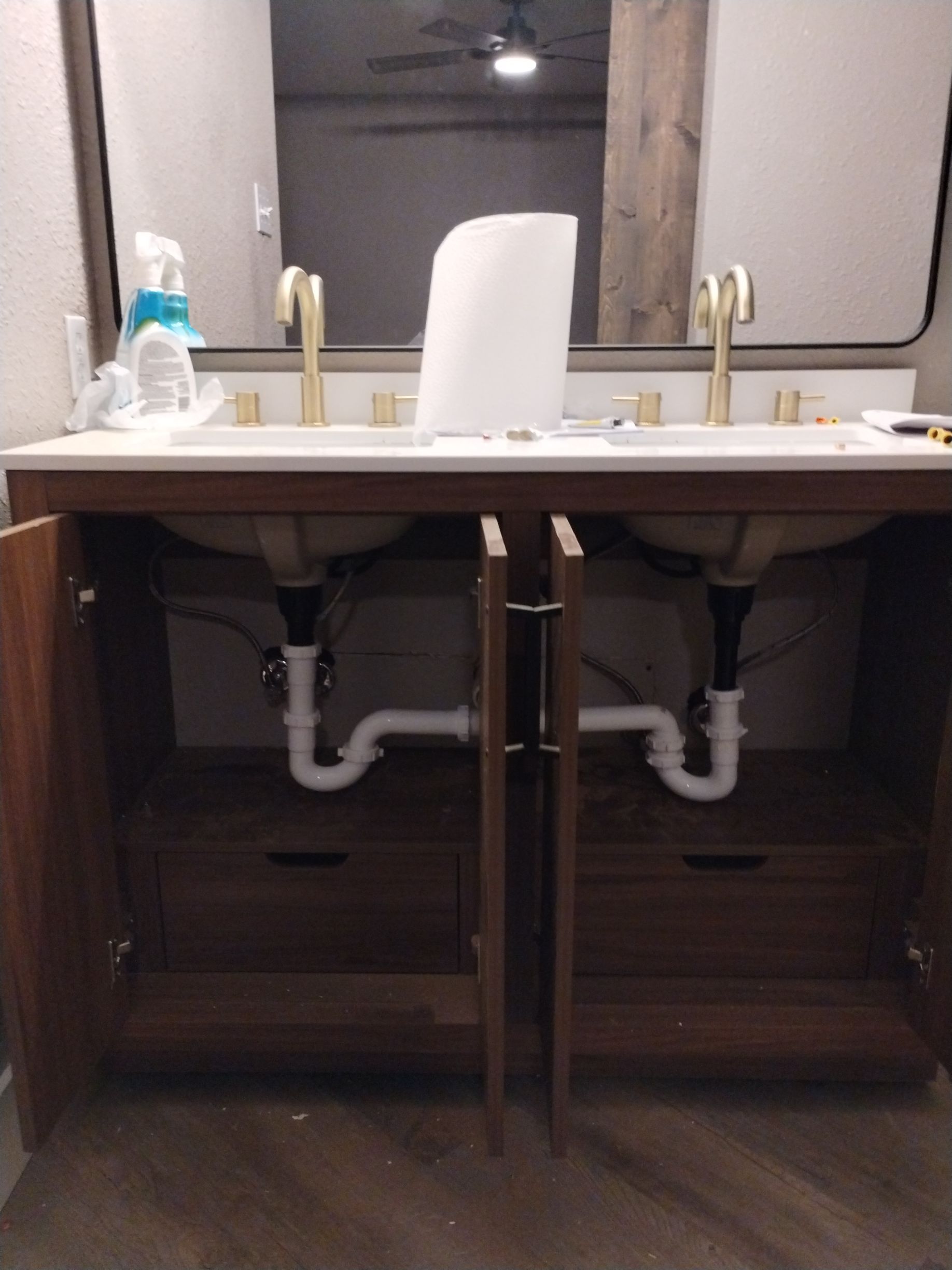 Bathroom vanity with two sinks, open cabinet doors, plumbing visible. Gold faucets. White countertop and sinks.