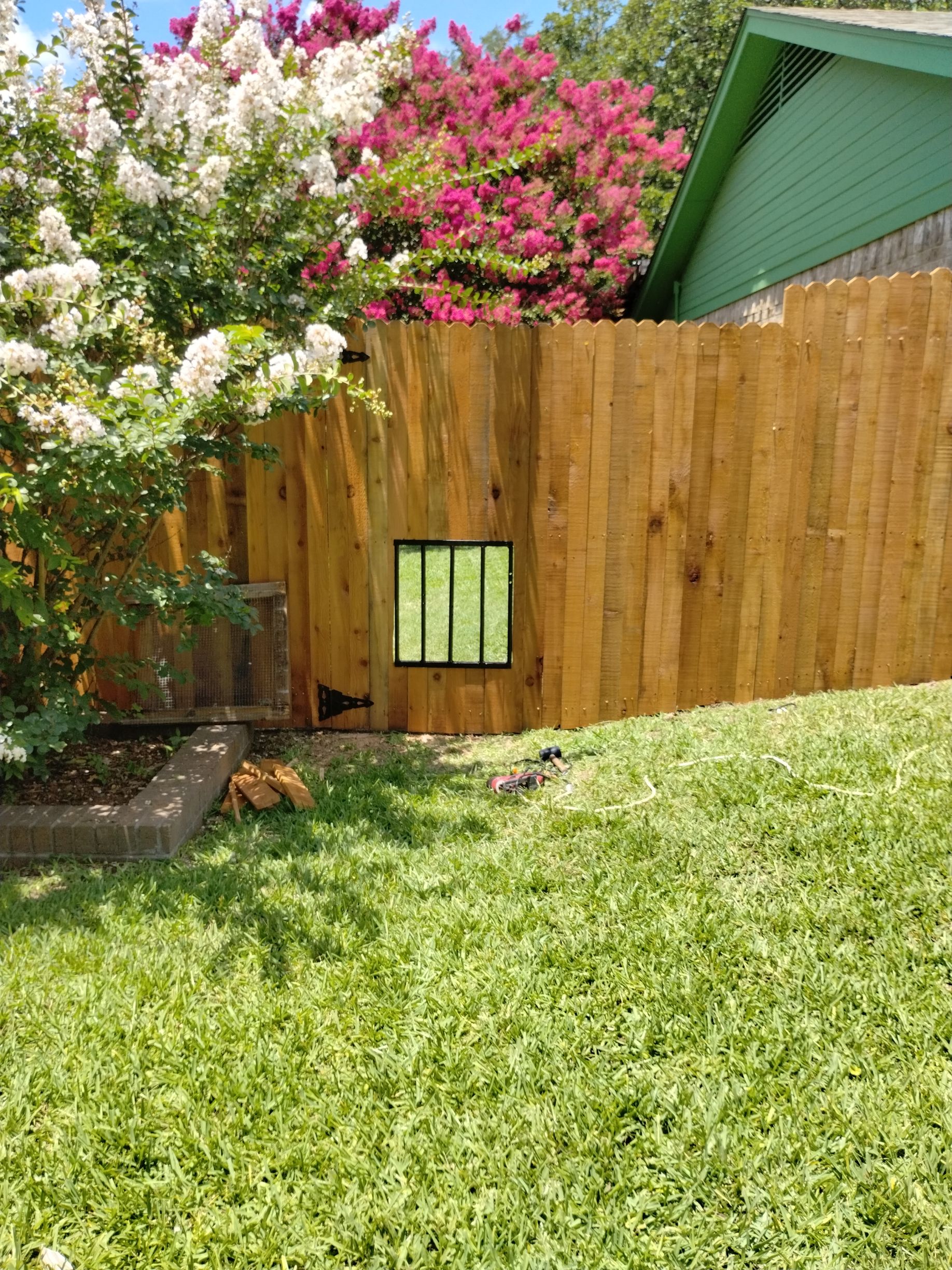 A wooden fence with a pet door. Colorful flowers and green grass surround it.