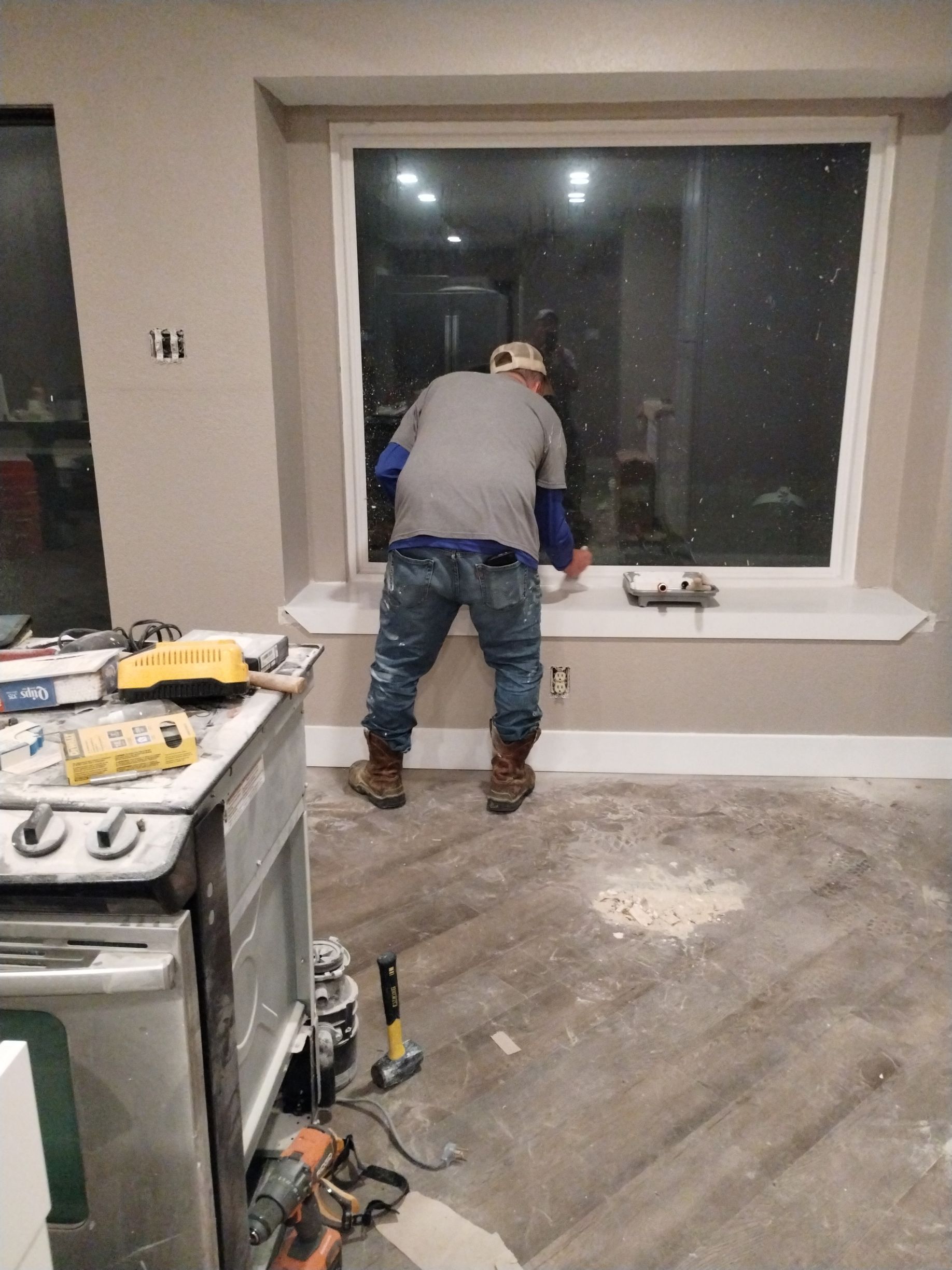 Man working near a large window in a room under construction; floor debris and tools visible.