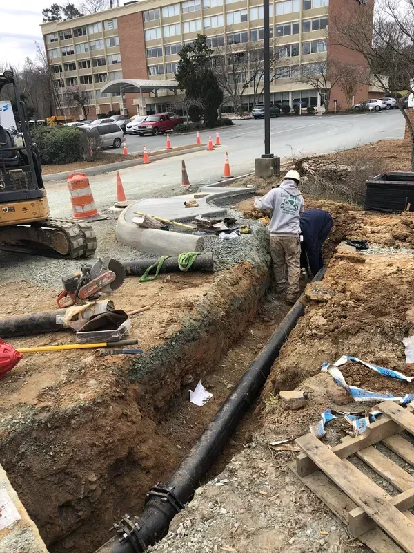 Construction workers installing a black pipe in an open trench near a building.