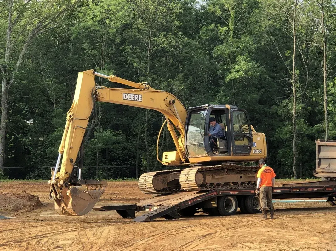 Yellow excavator on a trailer; worker in orange vest. Forest backdrop, sunny day.