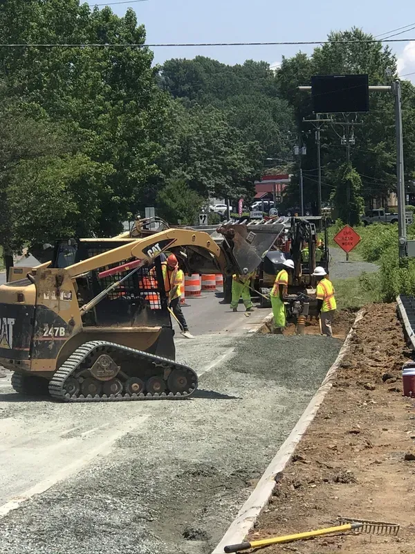 Construction workers near CAT skid steer compacting gravel on road. Orange cones and caution signs.