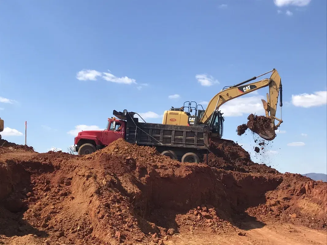 Excavator loading dirt into a red dump truck on a brown mound under a blue sky.