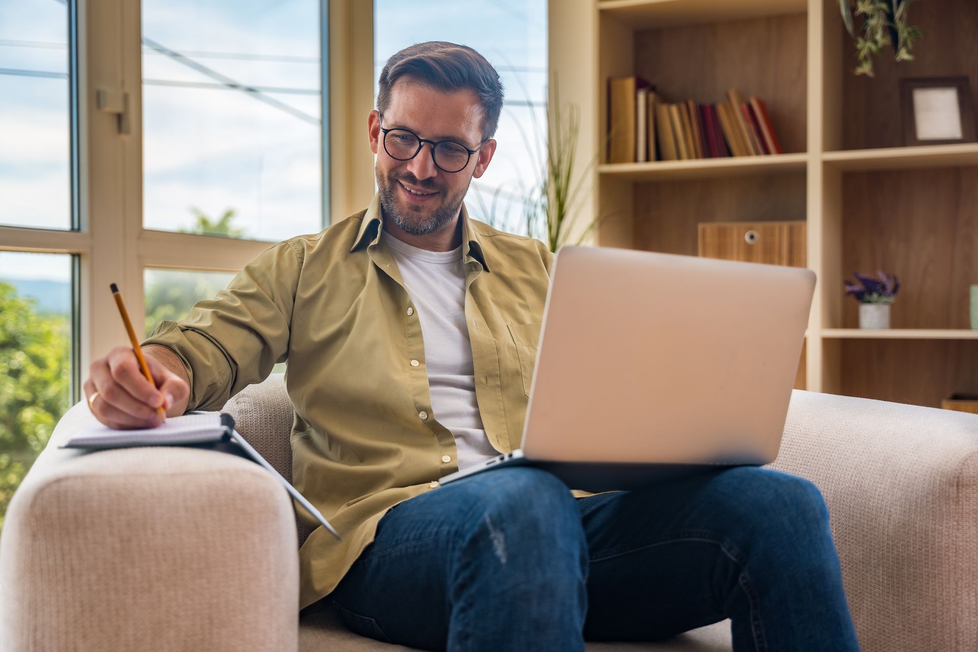 Man writing on a couch beside a laptop in a bright living room