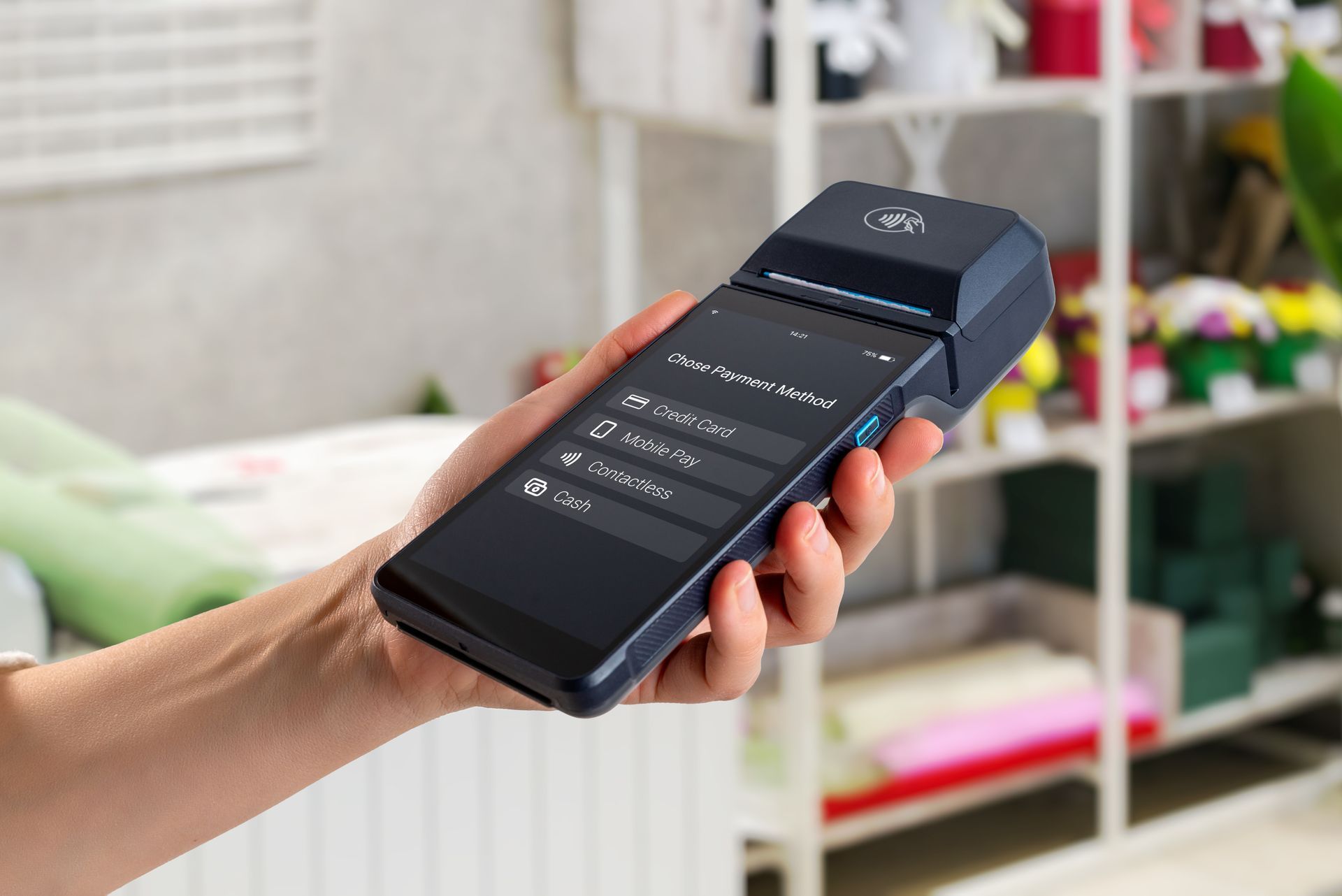 Hand holding a black touchscreen payment terminal with a menu on screen in a blurred retail setting