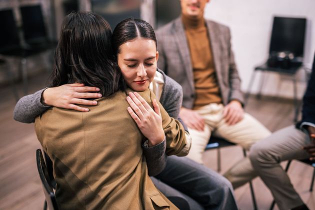 Two people sit close, hugging and comforting each other in a counseling group setting.