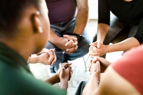 People seated in a circle, clasping hands in a group meeting or support session.