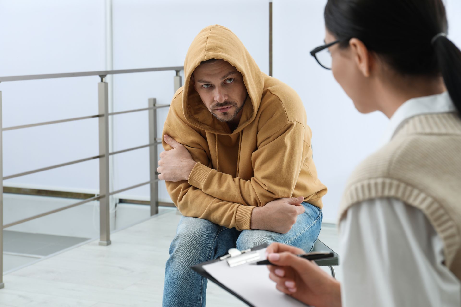 Man in a tan hoodie sits across from a clinician, leaning forward in a bright office.