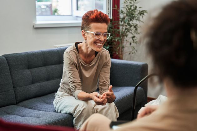 Two people talking on a gray couch in a bright living room, one smiling and leaning forward