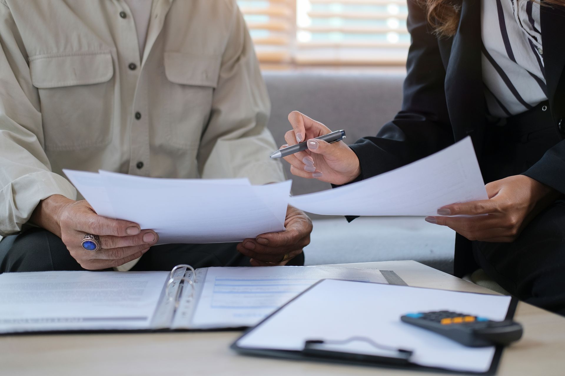 Two people reviewing papers at a desk with a binder and calculator in a bright office
