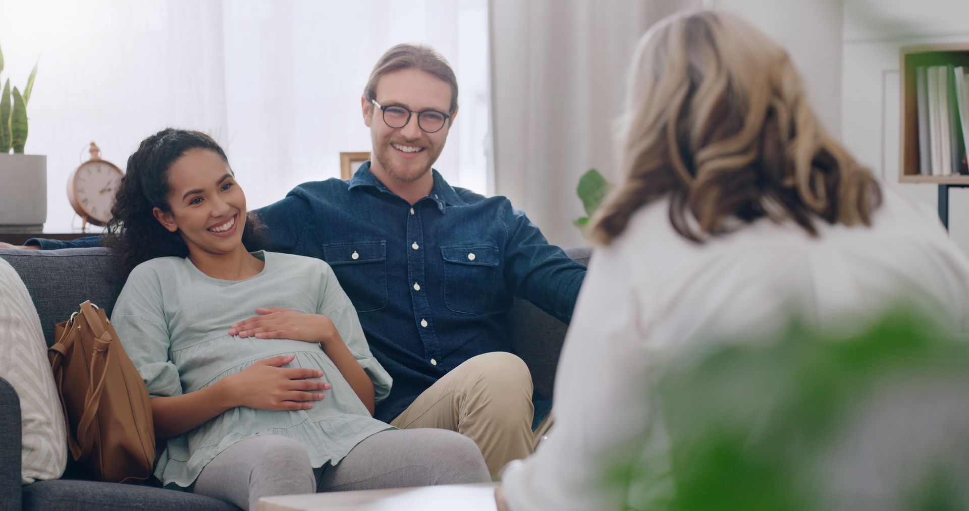 Pregnant woman and smiling man seated on a couch, talking with a counselor in a bright living room.