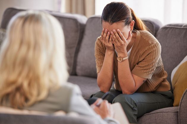 Woman sits on a couch with her face in her hands, appearing upset, while another person listens nearby.