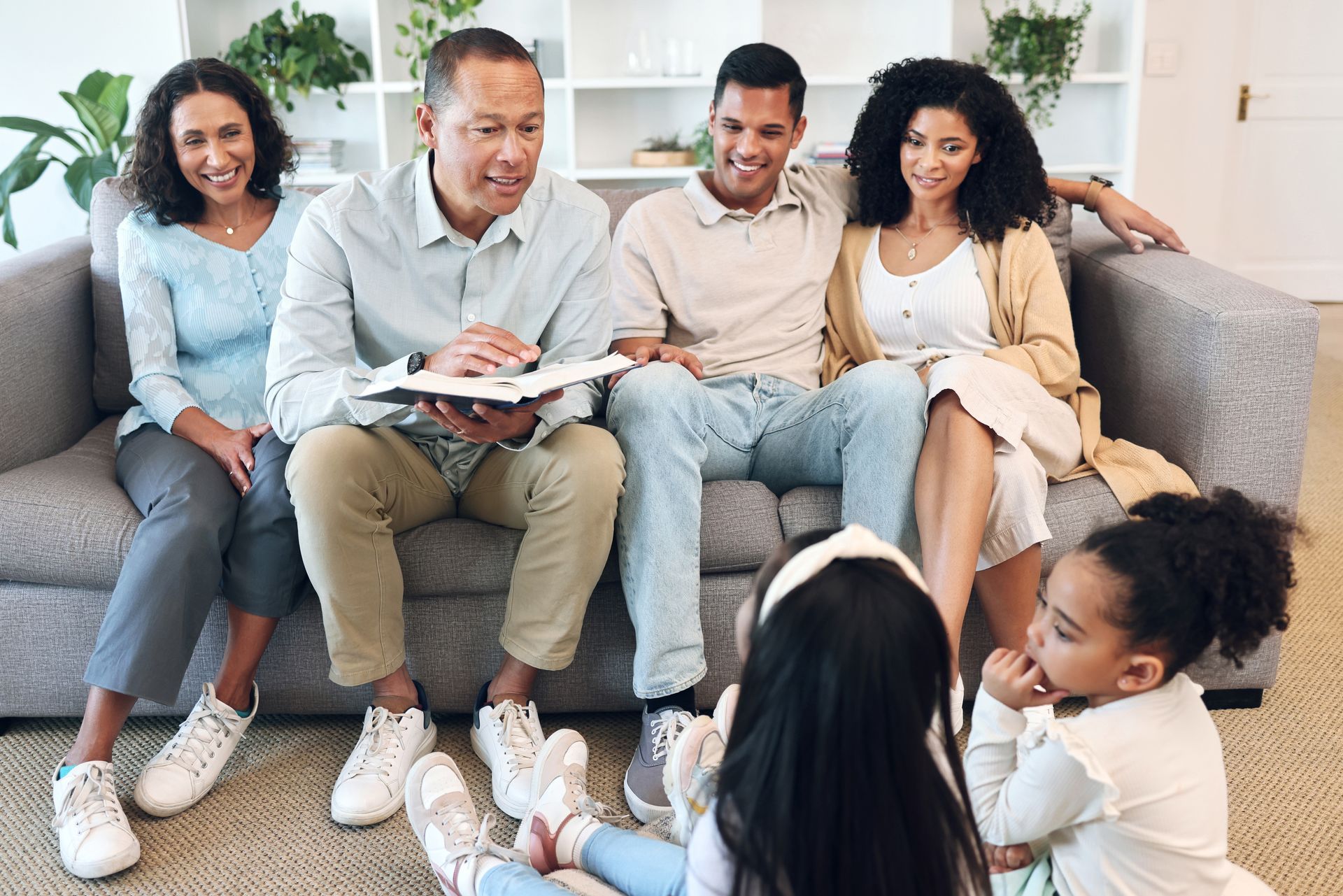 Four people on a couch reading from a tablet to children seated on the floor in a bright living room