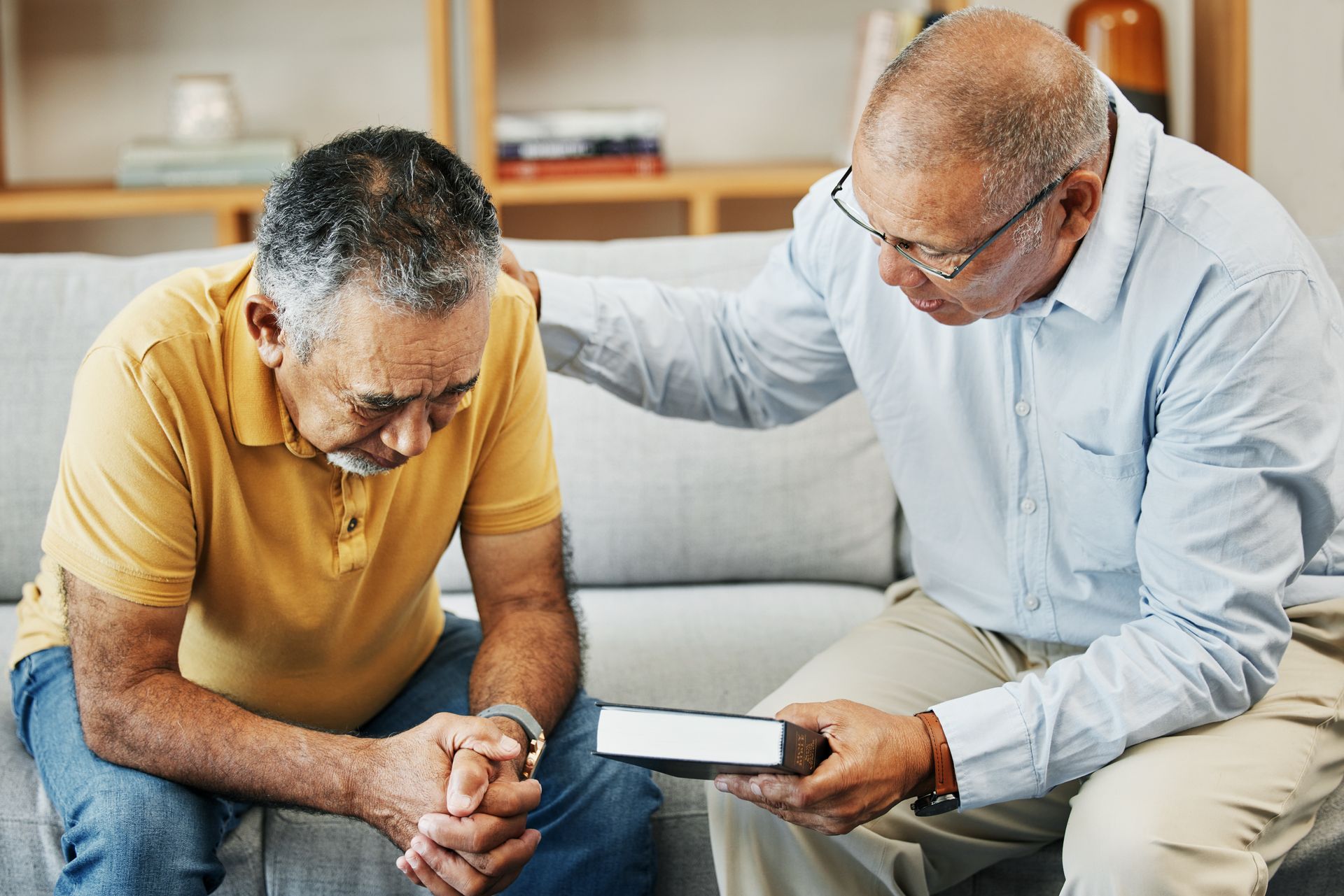 Two men sitting on a couch, one comforting the other while showing him a small book or device