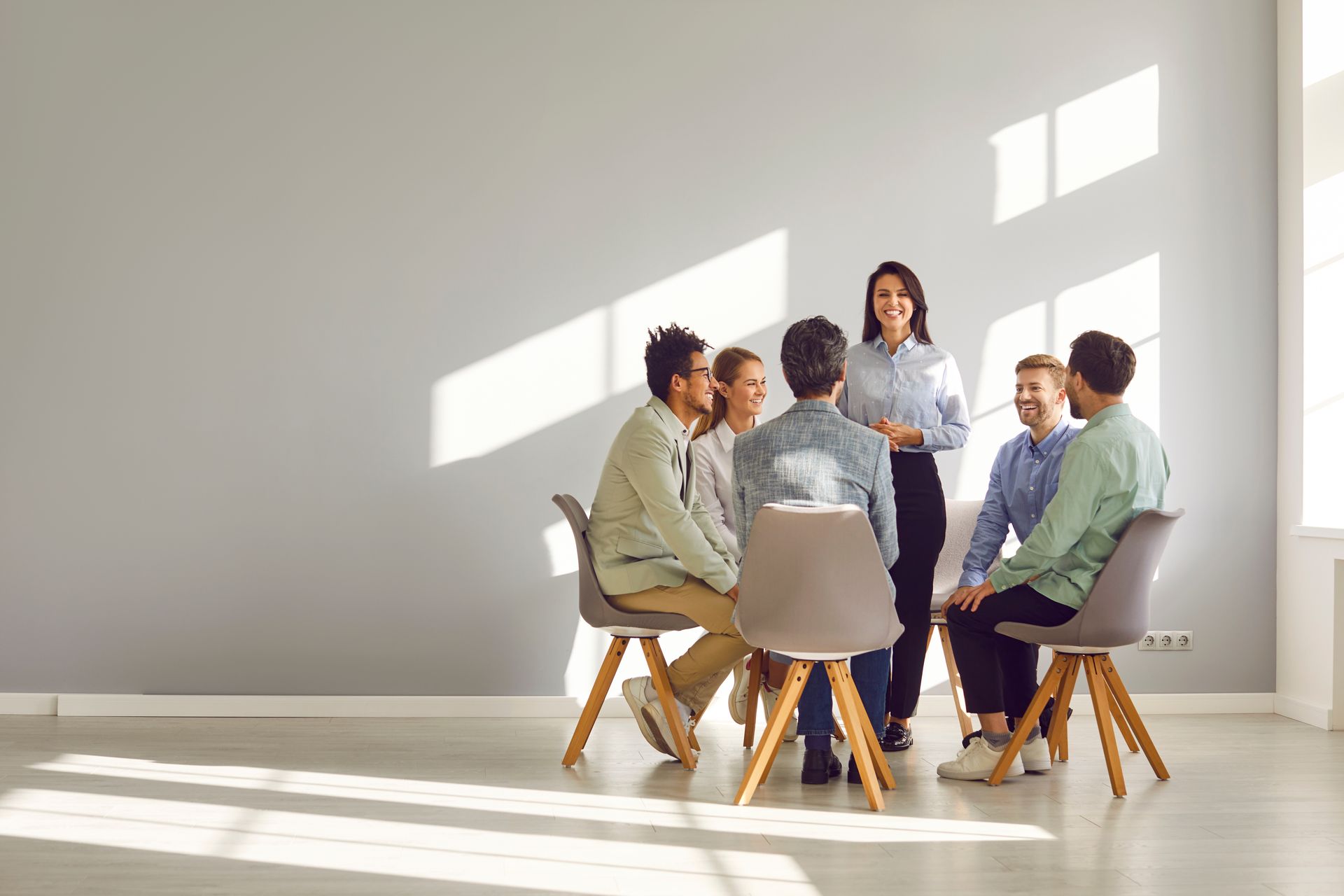 Five people sit in a bright meeting room, talking around a round table near a window.