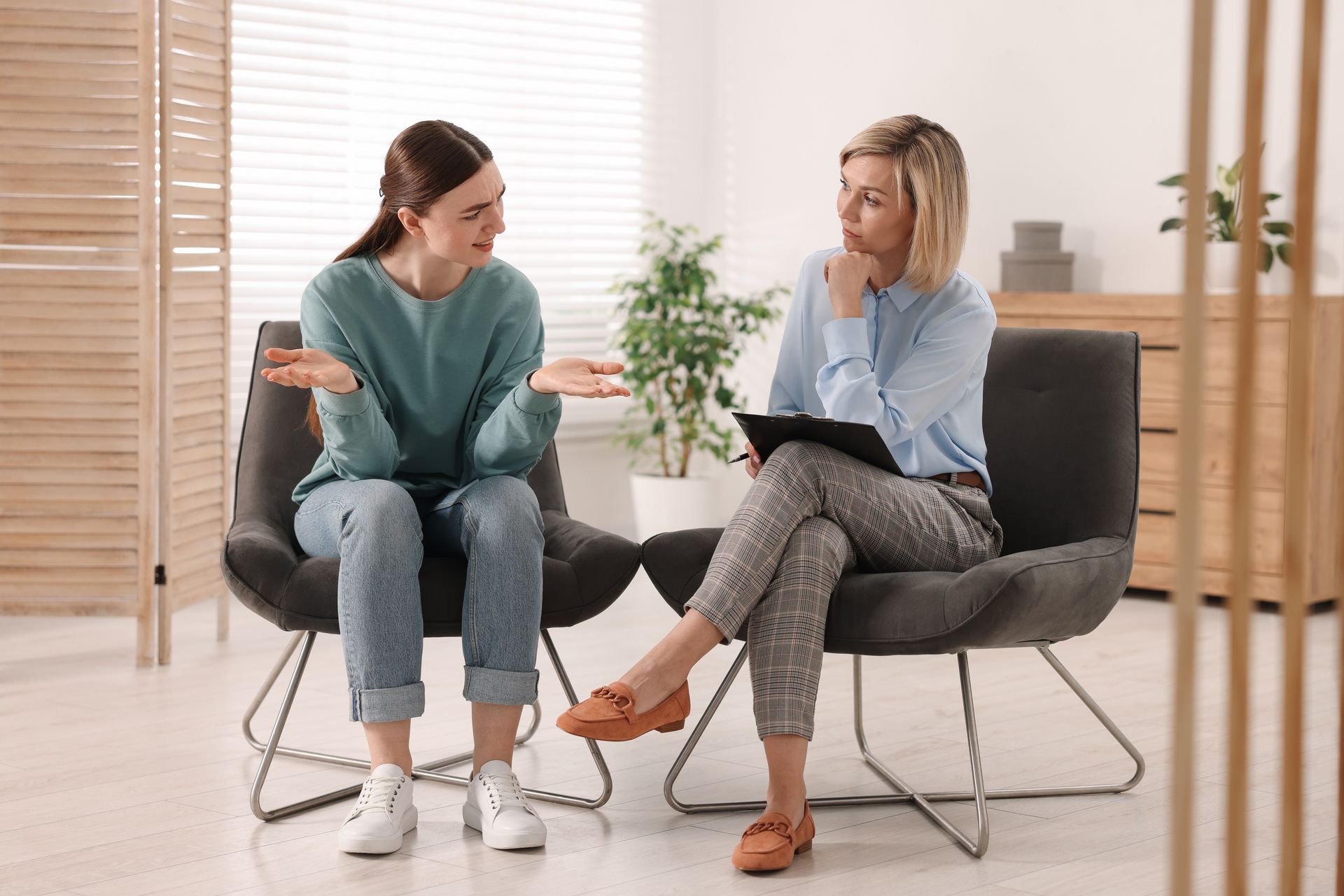 Two women talking in a bright living room, seated on modern chairs, one gesturing while the other listens.