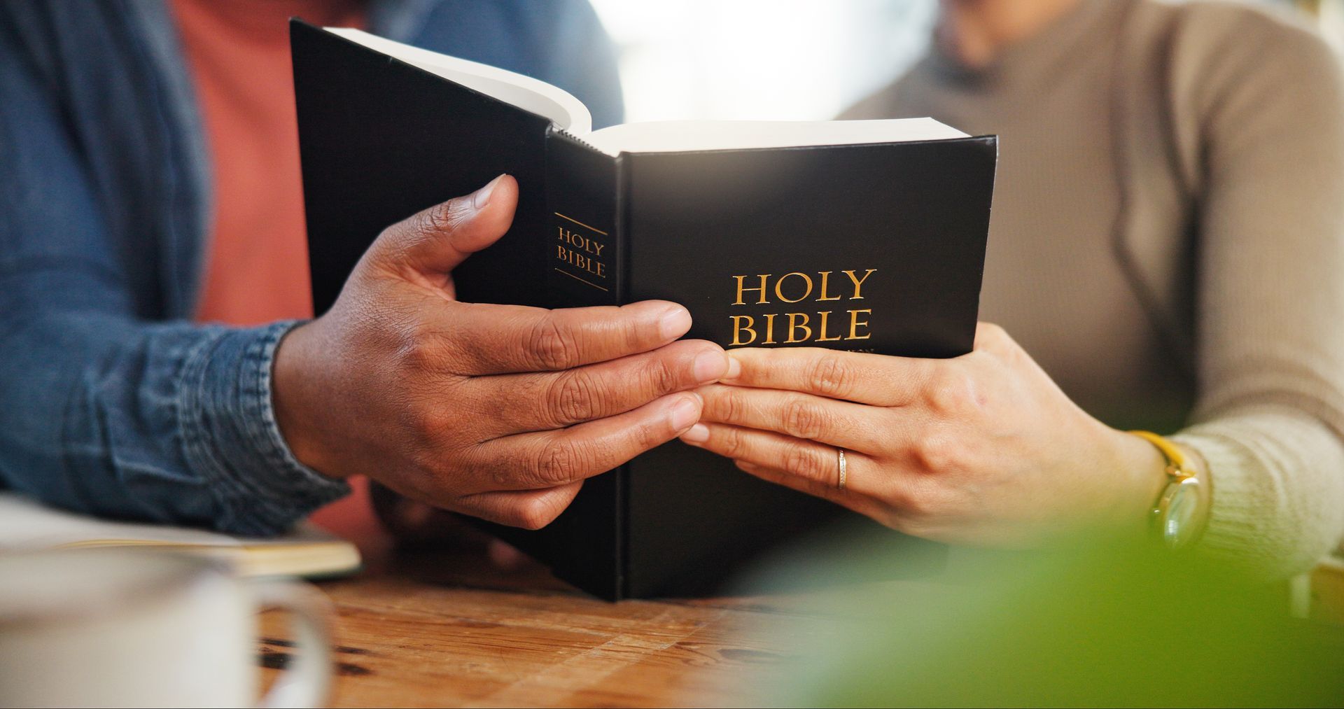 Hands holding a black Holy Bible on a wooden table, with a blue-shirted person nearby