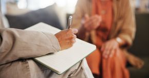 Person writing in a notebook during a conversation with another seated person indoors