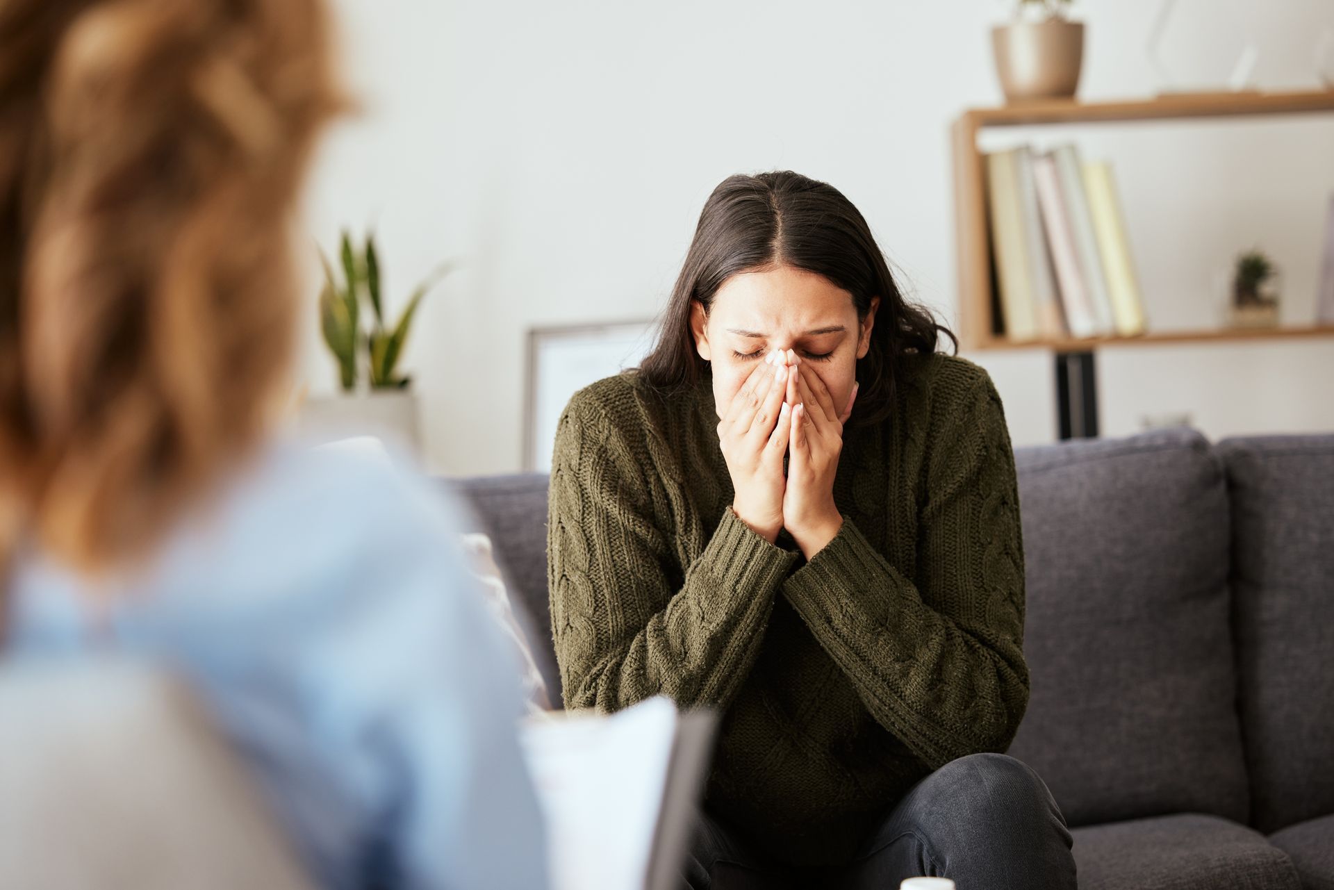 Woman sits on a couch with her face in her hands while talking to a therapist.