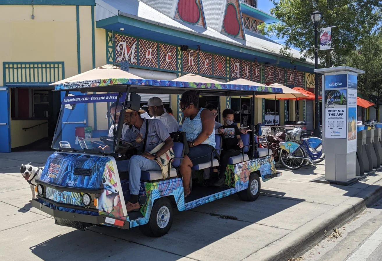 Colorful open-air tour tram parked on a sunny street beside a building and sidewalk.