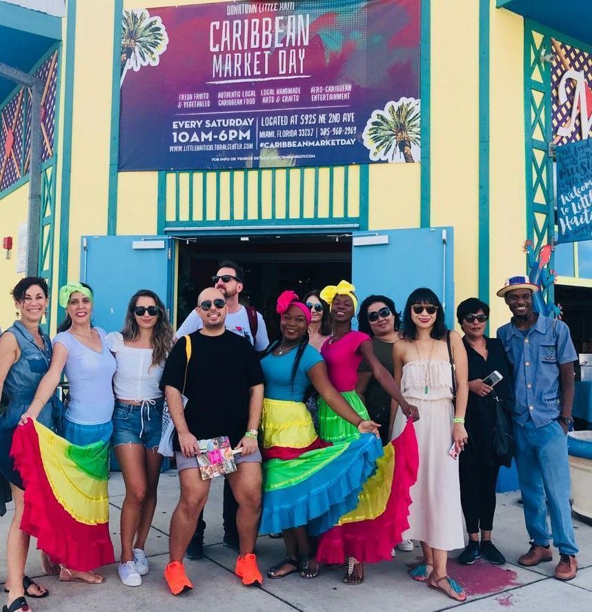 People posing outside a Caribbean Market storefront, many wearing bright rainbow skirts and festive clothing