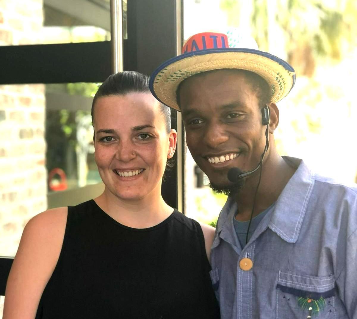 Two smiling people posing together outdoors, one in a black top and one wearing a straw hat with a headset