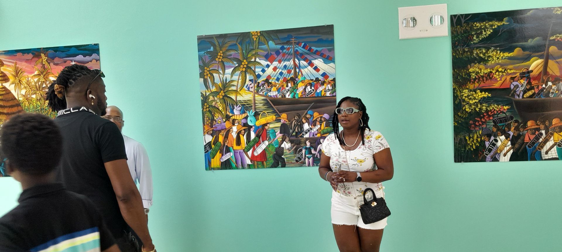 People viewing colorful paintings in a teal-walled gallery, with a woman speaking near the center.