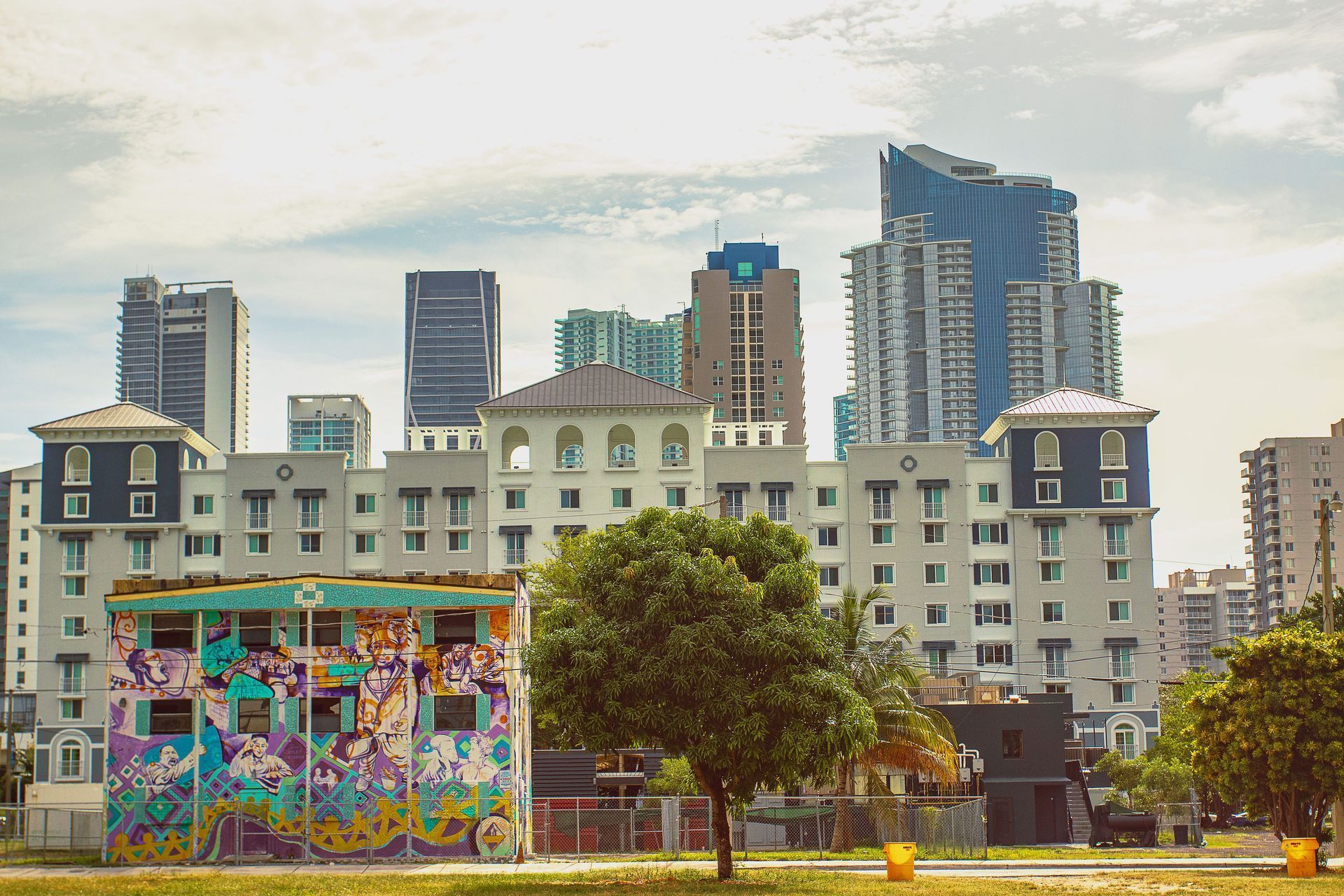 City skyline behind a low building with colorful mural and a large tree in the foreground.