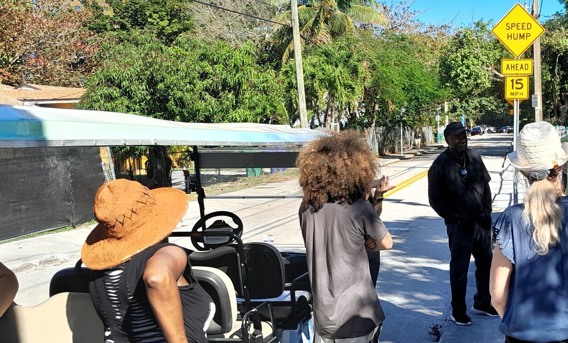 People gathered near a golf cart on a sunny road by a yellow caution sign.